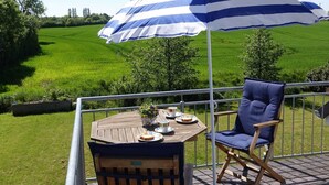 A wooden deck with a table, a blue and white striped umbrella, and a blue chair overlooking a green field. - Ferienwohnung Regenbogenhaus - Ferienwohnung Regenbogenhaus: Special Rate May and June! (Hasselberg)