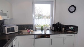A modern kitchen with white cabinets, a black countertop, a microwave, a coffee maker, and a clock on the wall. - Ferienwohnung Regenbogenhaus - Ferienwohnung Regenbogenhaus: Special Rate May and June! (Hasselberg)