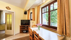 Una habitación con televisión sobre un mueble bajo, una mesa de comedor con sillas, un ventanal con cortinas y una puerta con panel de vidrio.