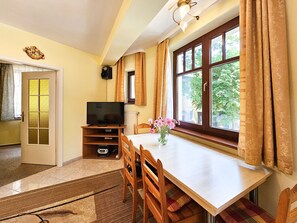 Une pièce comprenant un téléviseur posé sur un meuble, une table à manger avec des chaises, une fenêtre avec des rideaux et une porte avec un panneau de verre.