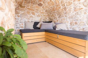 A corner sofa with cushions against a stone wall, wooden drawers, and a tiled floor.