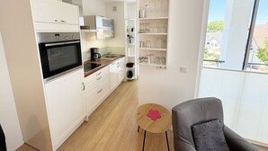 A compact kitchen with white cabinets, a built-in oven, and a coffee maker. There's a small round table with a red flower decoration. - A: Villa Antje Whg. 12 with balcony - Villa Antje Whg. 12 with balcony (Göhren)