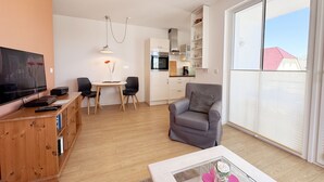 A modern living room with a flat-screen TV, wooden furniture, a grey armchair, and a large window offering a view of the outside. - A: Villa Antje Whg. 12 with balcony - Villa Antje Whg. 12 with balcony (Göhren)
