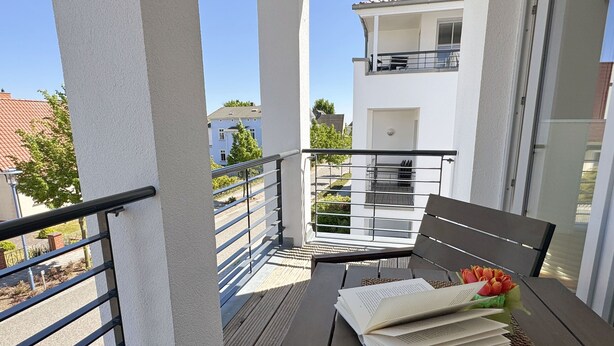 A balcony with a wooden bench, a table with books and a bouquet of flowers, and a view of neighboring buildings. - A: Villa Antje Whg. 12 with balcony - Villa Antje Whg. 12 with balcony (Göhren)