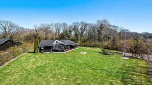 Ein modernes Haus mit großem Rasen, einer überdachten Sitzgelegenheit im Freien und Blick auf eine bewaldete Landschaft.