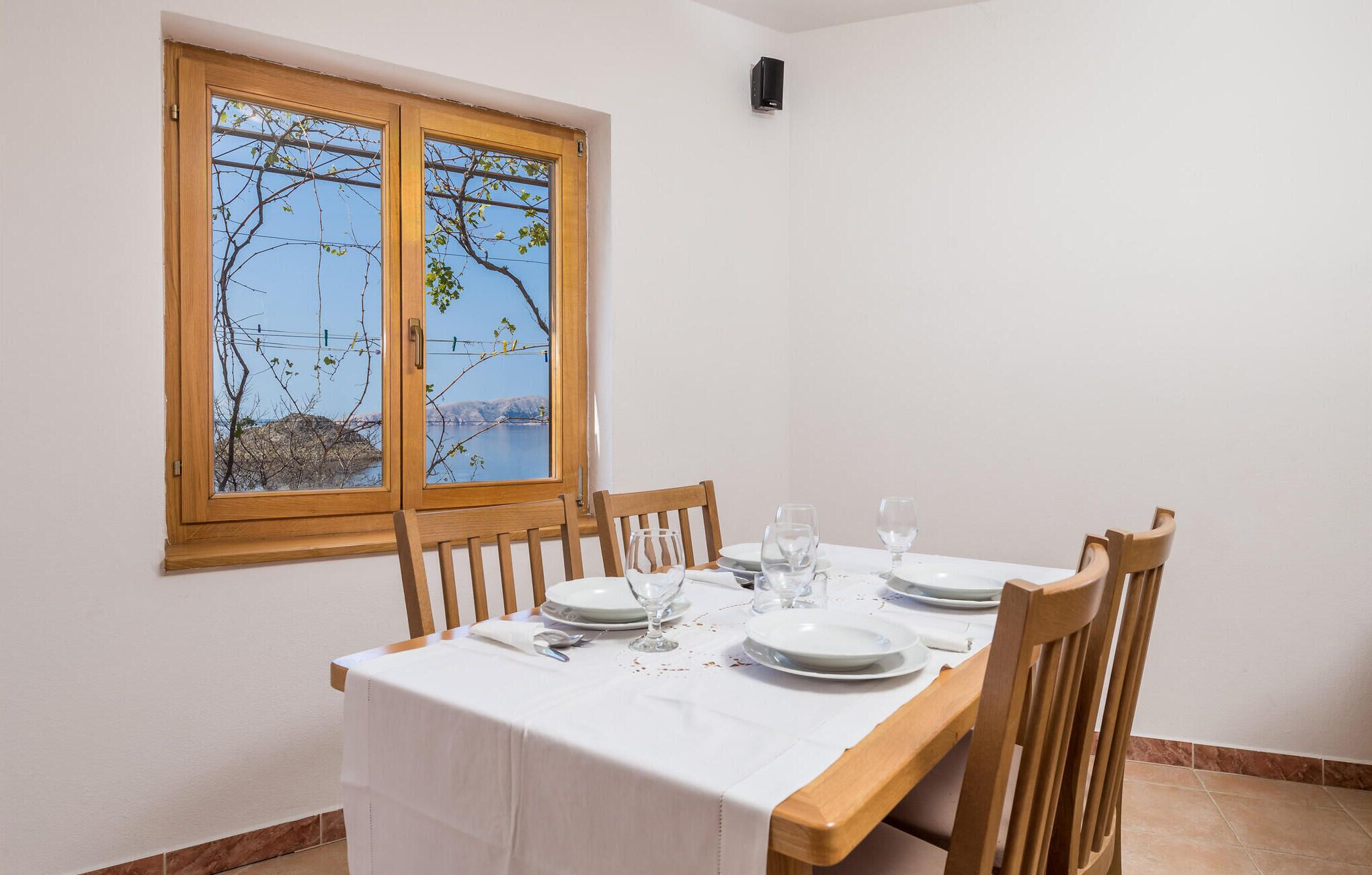 A dining area with a table set for four, a window with a view of a body of water and hills, and a wall-mounted air conditioning unit.