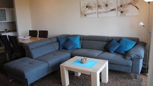 Ein modernes Wohnzimmer mit einem grauen Ecksofa, einem kleinen Holztisch und einem Essbereich mit Tisch und Stühlen.