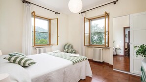 Quarto com cama, cadeira, duas janelas com cortinas, radiador e vista para o verde lá fora.