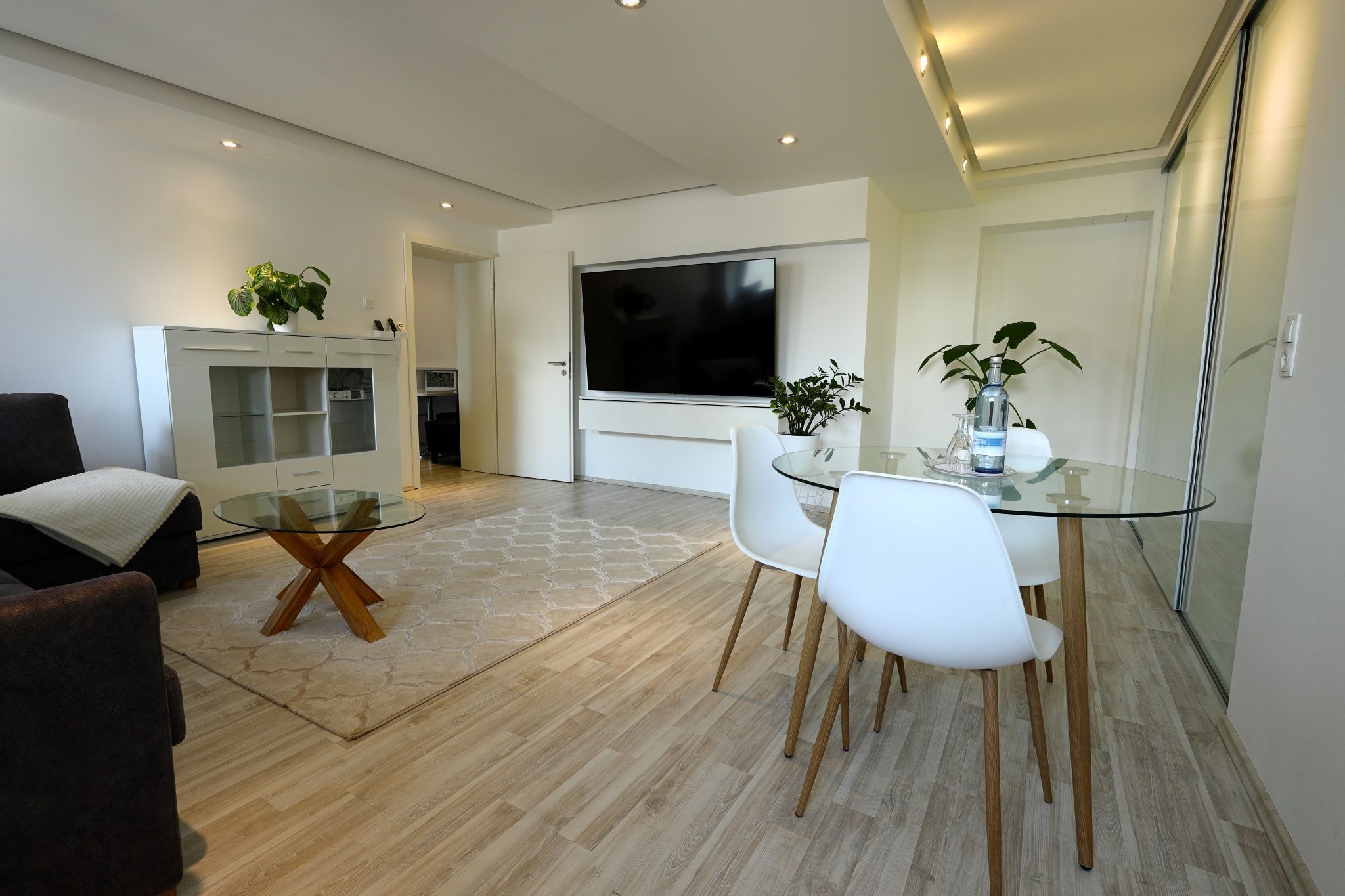 A modern living room with a glass dining table, white chairs, a sofa, and a TV mounted on the wall.