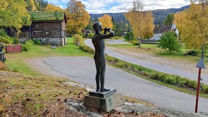 Une statue représentant une personne, placée sur un piédestal, dans un cadre rural avec des maisons et des arbres.