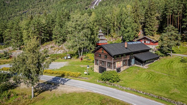 Une cabane en bois, dotée d’un toit à forte pente, entourée d’une forêt dense et avec une chaîne de montagnes en arrière-plan.