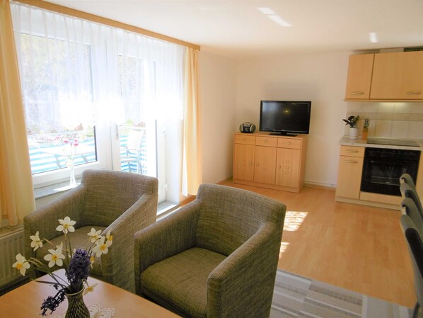 A living room with two green armchairs, a small table with a vase of flowers, a TV on a wooden cabinet, and a kitchen area with wooden cabinets and a stove.