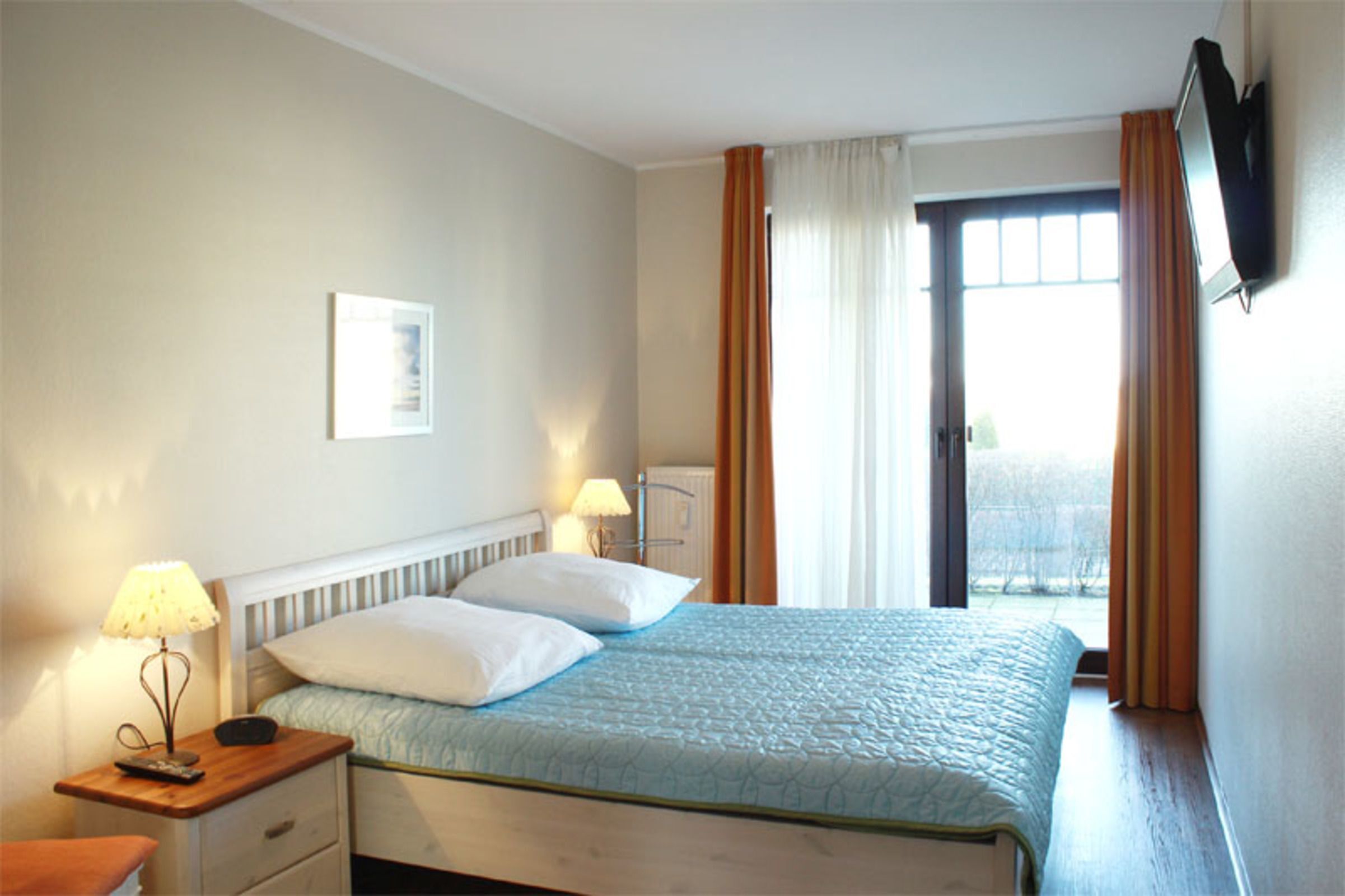 Un dormitorio con una cama, dos mesitas de noche, una lámpara, un televisor montado en la pared y una ventana con cortinas.