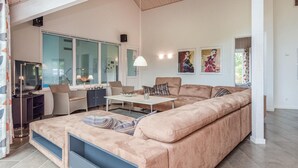 Ein modernes Wohnzimmer mit einem großen, beigefarbenen Ecksofa, einem kleinen Tisch und zwei Sesseln. Ein großes Fenster bietet einen Blick ins Freie, und an der Wand hängt ein Gemälde.