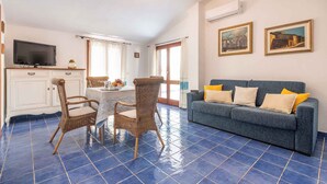 A living room with a blue tiled floor, a blue sofa with yellow cushions, wicker chairs, a dining table, and a TV on a cabinet. - Holiday apartment mit Meerblick (Cala Gonone)