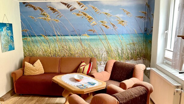 A cozy living room with a large beach mural, a brown sofa, a small table, and a window with a view of the sea. - Rügen-apartment 65 - apartment (Garz/Rügen)