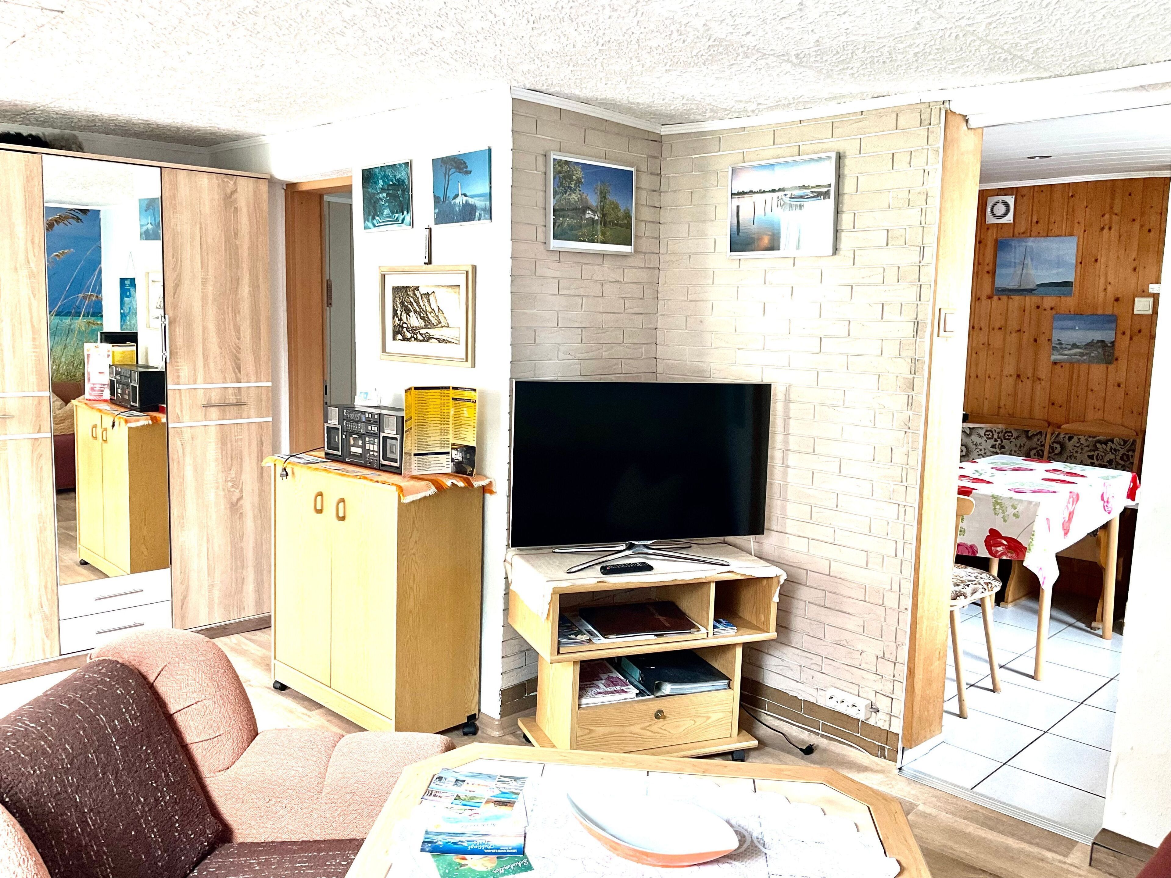 Un espacio habitable compacto con un televisor sobre un mueble de madera, una pequeña mesa de comedor con mantel floral y un sofá.
