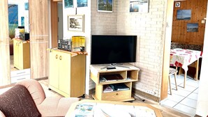 A compact living space with a TV on a wooden stand, a small dining table with a floral tablecloth, and a sofa. - Rügen-apartment 65 - apartment (Garz/Rügen)