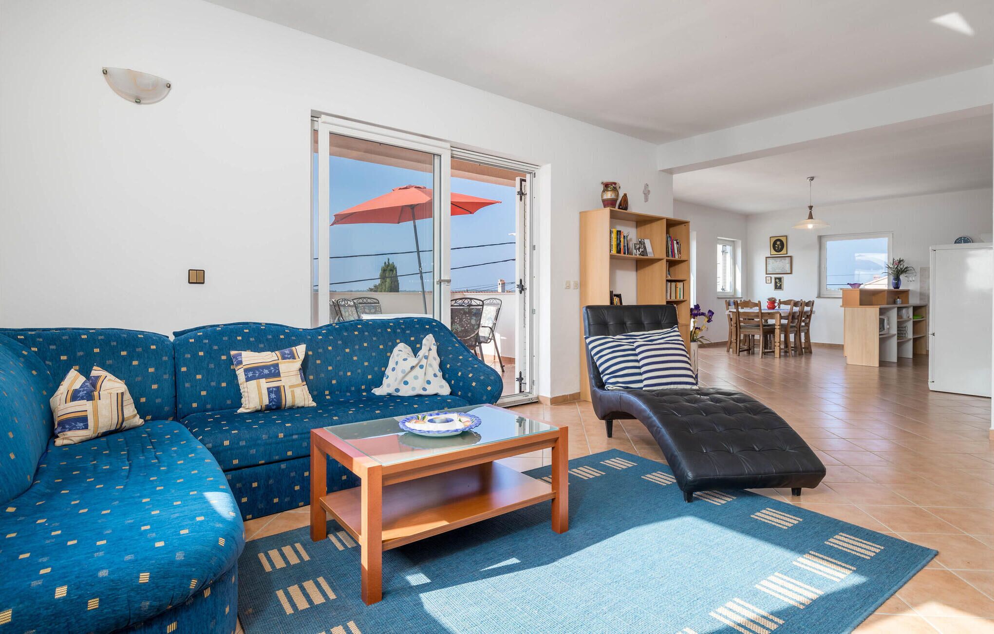A living room with a blue sofa, a wooden coffee table, and a black ottoman. There is a sliding glass door leading to a balcony with a red umbrella.
