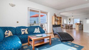 Un salon avec un canapé bleu, une table basse en bois et un pouf noir. Une porte-fenêtre coulissante donne accès à un balcon agrémenté d’un parasol rouge.