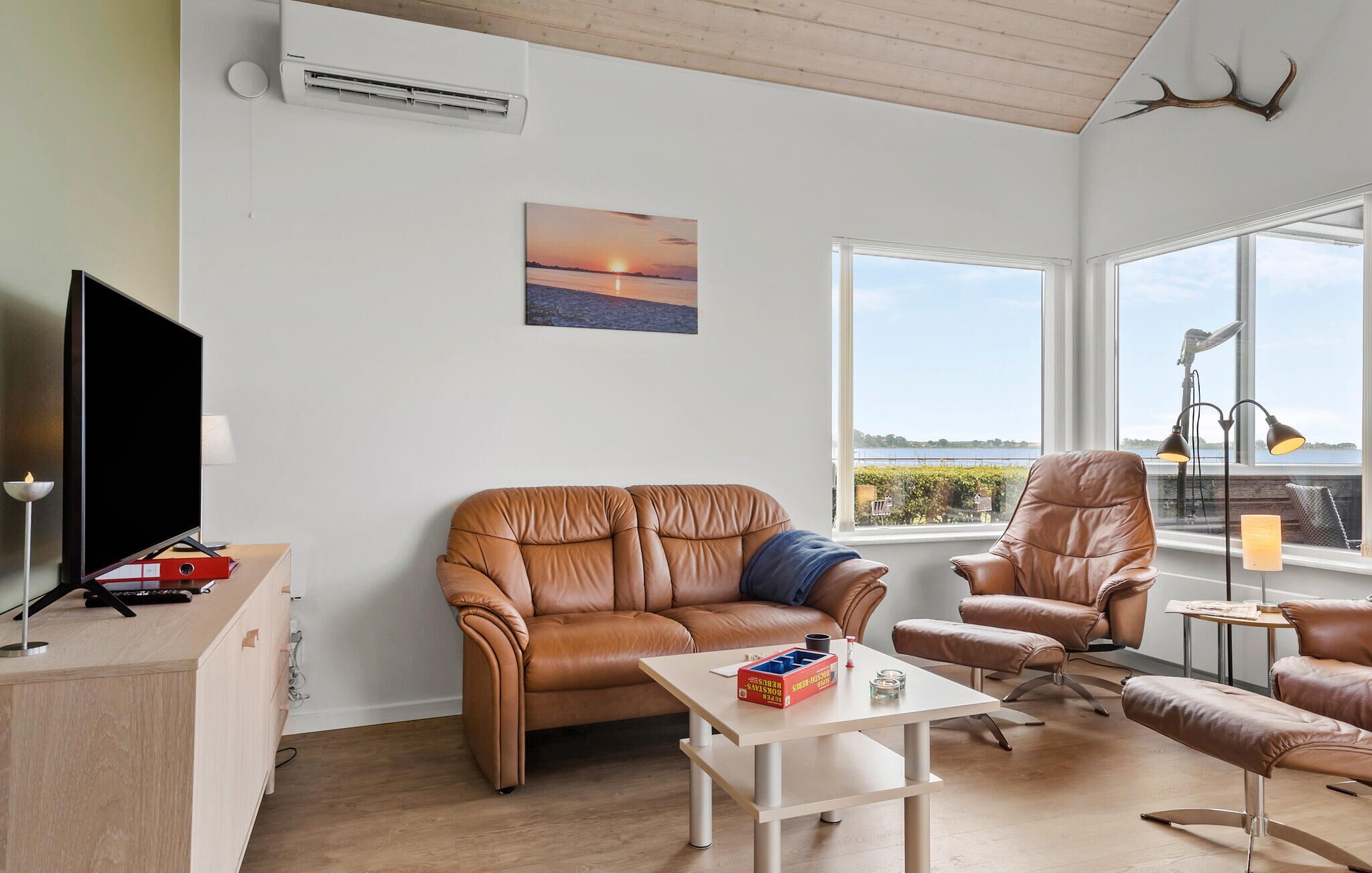 Un salon moderne avec un canapé en cuir, une table basse et un téléviseur. On y trouve un tableau représentant un coucher de soleil, une grande fenêtre offrant une belle vue et une décoration murale en forme de bois de cerf.