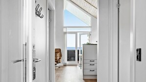 A hallway with a white door, a coat rack, a leather chair, a desk with drawers, and a view of the sky through a glass door.
