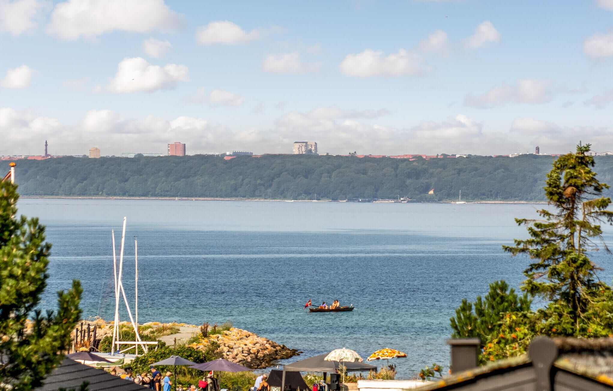 Blick auf die Küste mit ruhiger See, einem Schiffen im Hintergrund und einem bewaldeten Hügel.
