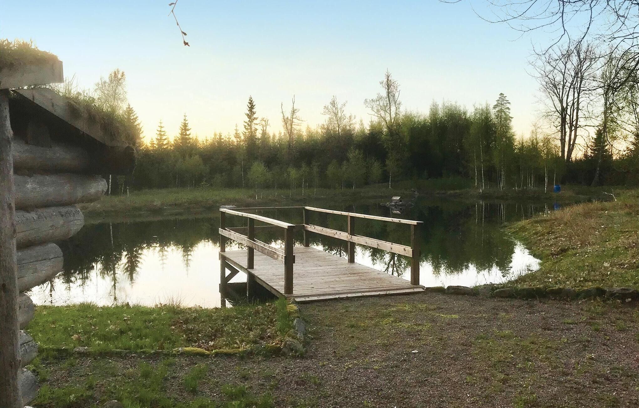 Un muelle de madera se extiende sobre un lago tranquilo, rodeado de un bosque denso y una cabaña de troncos.