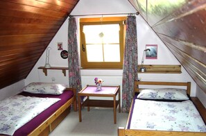 A small, cozy attic bedroom with two single beds, a wooden table, and a window with curtains.
