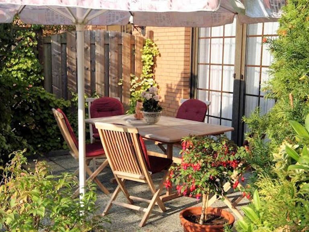 Um pátio com mesa e cadeiras de madeira, rodeado por plantas em vasos e uma parede de tijolos.