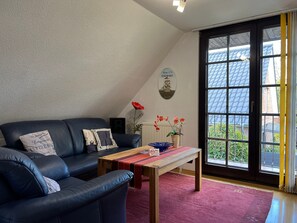 A living room with a black leather sofa, a wooden coffee table, and a red rug. There is a window with a view of a blue-roofed building outside.