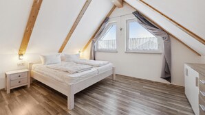 A bedroom with a sloped ceiling, a bed with white bedding, a nightstand, a dresser, and two windows with curtains. - Ferienhaus 2 Altefähr (Altefähr/Rügen)