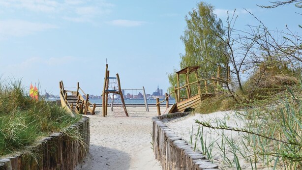 A sandy beach path leading to a wooden structure with a rope ladder, surrounded by grass and trees, with a cityscape in the background. - Ferienhaus 2 Altefähr (Altefähr/Rügen)