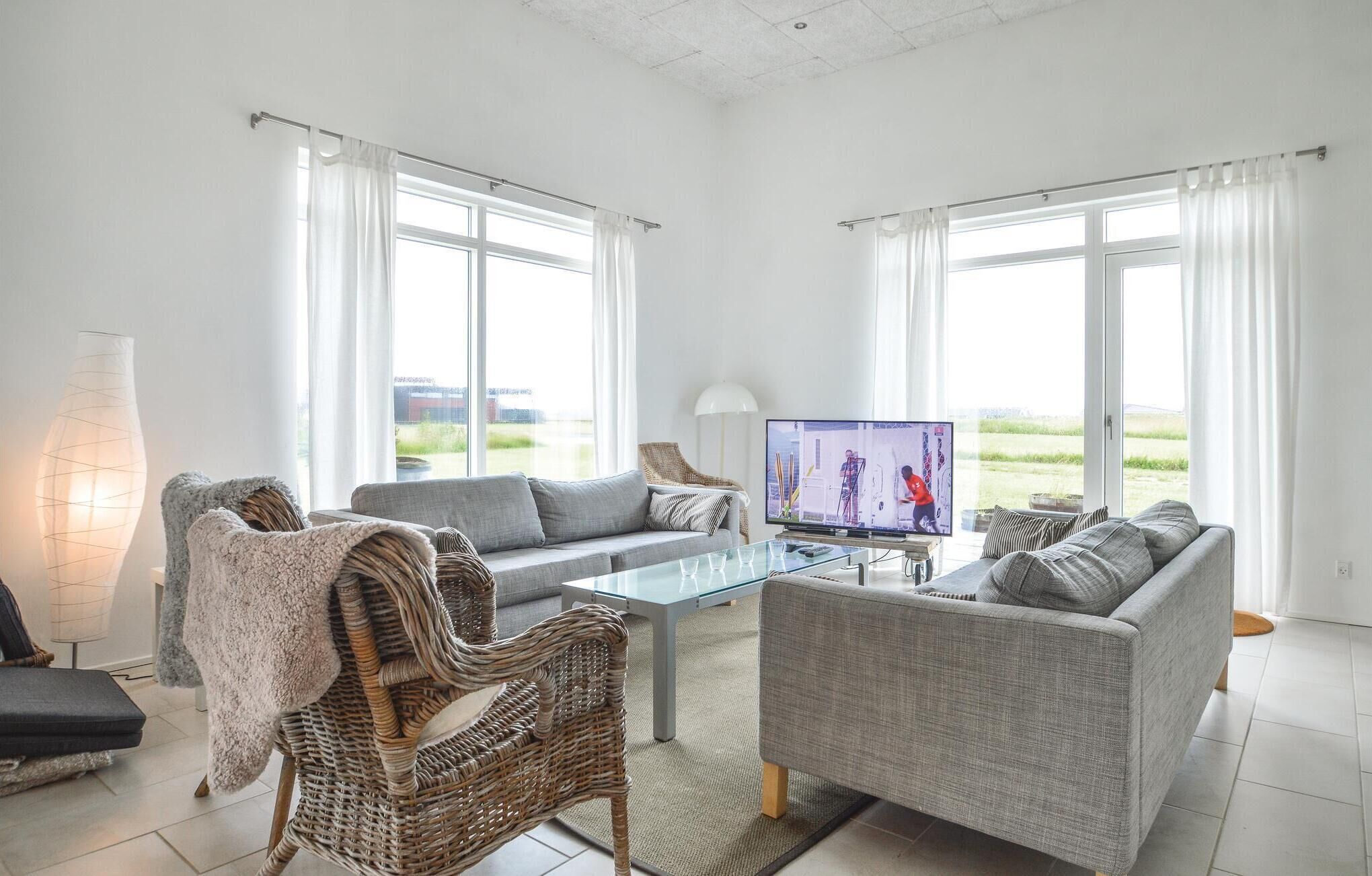 Ein modernes Wohnzimmer mit zwei grauen Sofas, einem Glastisch, einem Korbstuhl und einem Fernseher, der ein Sportspiel zeigt.