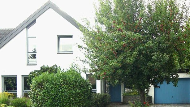 Una casa blanca con puerta de garaje azul, rodeada de vegetación y un camino de adoquines.