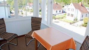 Quarto com uma mesa e duas cadeiras de vime, grandes janelas com vista para casas.