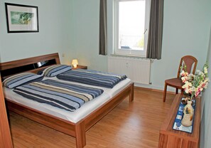 Une chambre à coucher avec un lit en bois, une table de chevet, une chaise, une fenêtre avec des rideaux et un vase rempli de fleurs.