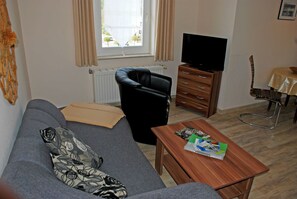 Un salon comprenant un canapé, une table basse en bois avec des magazines, un fauteuil en cuir noir, un meuble en bois avec un téléviseur et une fenêtre avec des rideaux.