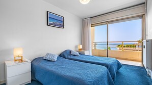 Ett hotellrum med två sängar, ett nattduksbord med en lampa, en inramad tavla på väggen och en balkong med utsikt över havet.