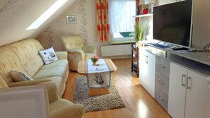 Ein gemütliches Wohnzimmer mit Sofa, Sessel und Couchtisch, einem Fernseher und einer Küchenzeile mit Schränken und einem Heizkörper.