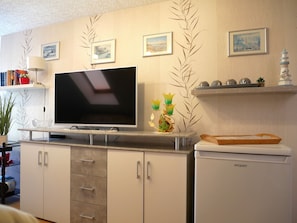 Ein modernes Wohnzimmer mit einem Flachbildfernseher auf einem Ständer, einer weißen Kommode mit Schubladen, einem kleinen Kühlschrank und Dekorationselementen auf Regalen.