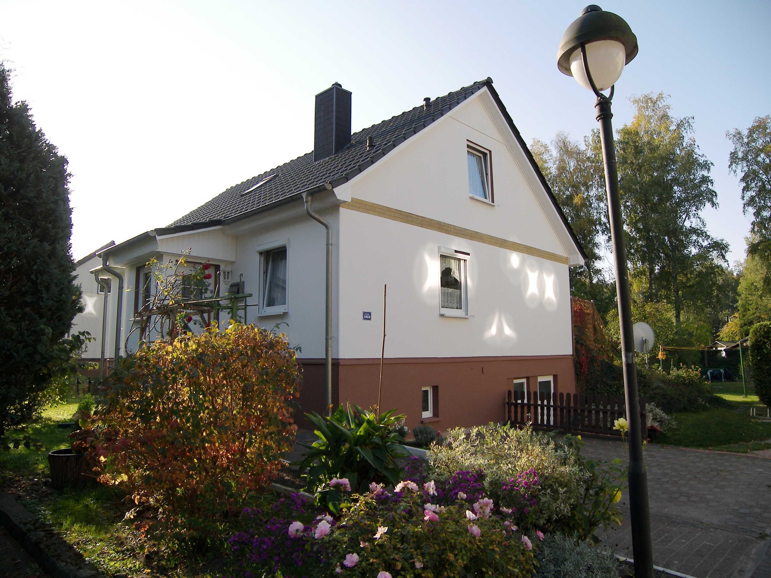 Ein zweistöckiges Haus mit weiß-brauner Fassade, einem sichtbaren Schornstein und einem Garten mit verschiedenen Blumen und Sträuchern.