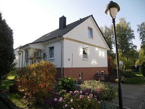 Une maison à deux étages, à l’extérieur blanc et marron, avec une cheminée bien visible et un jardin où poussent différentes sortes de fleurs et d’arbustes.