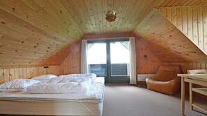 Une chambre mansardée chaleureuse avec un plafond en pente en bois, un lit avec une literie blanche, un fauteuil en cuir et une petite table.