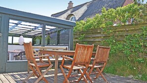 Une terrasse en bois avec une table et des chaises, une véranda en verre et une vue sur un bâtiment dont le toit est recouvert de chaume.