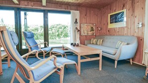 Une pièce avec un intérieur en bois, un canapé gris, deux fauteuils bleus, une table en bois et une grande fenêtre avec des rideaux.