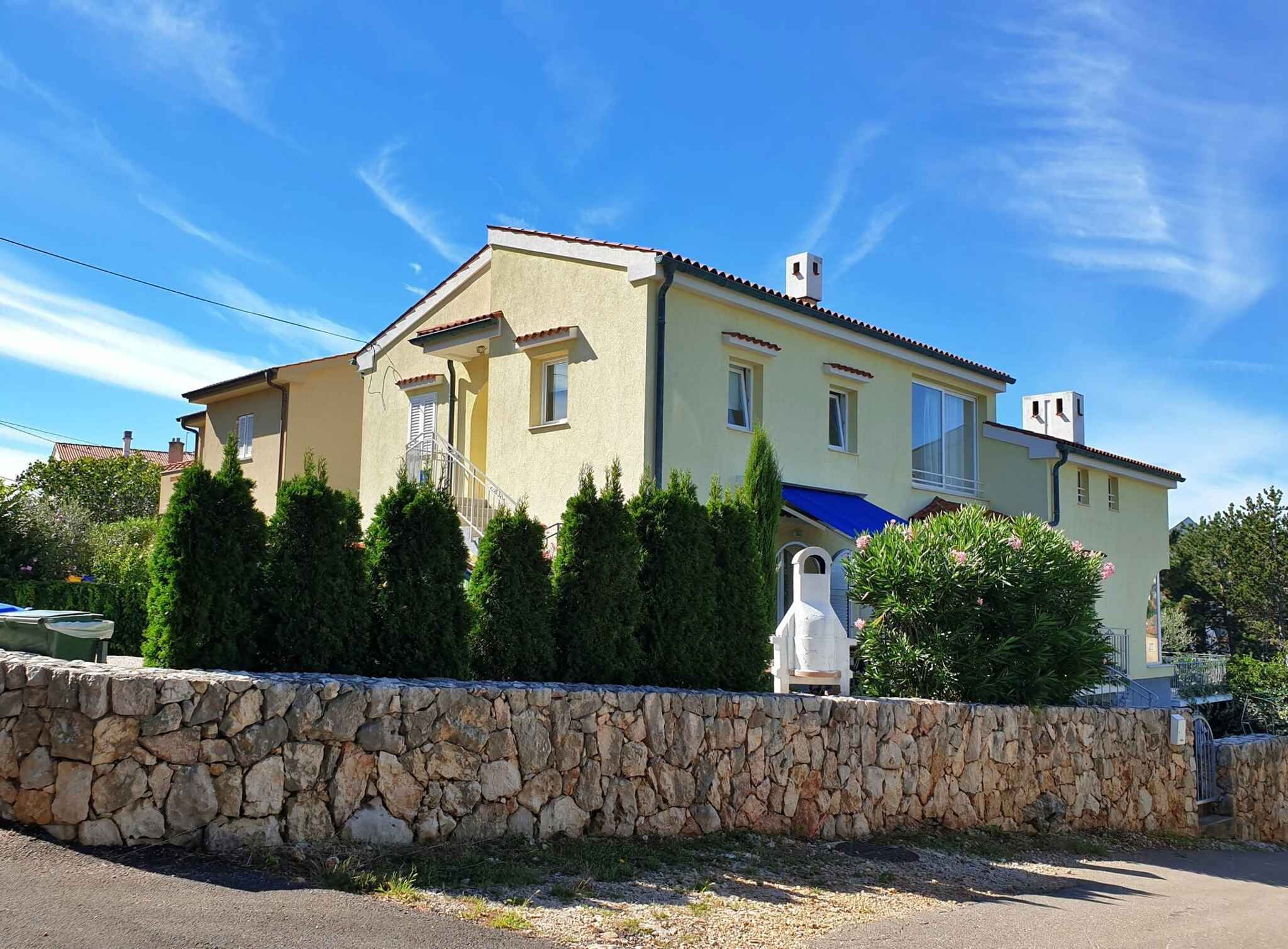 Een huis met twee verdiepingen, een stenen muur ervoor, een blauw zonnescherm en een heldere blauwe lucht.