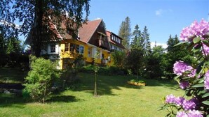 Une maison jaune avec un toit rouge, entourée d’un jardin bien entretenu et d’arbres.