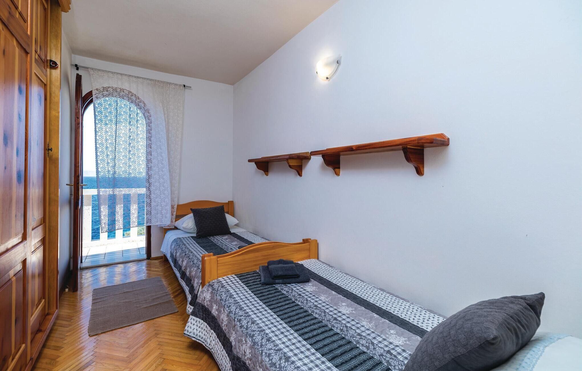 A bedroom with two single beds, wooden headboards, a small wooden shelf, and a view of the sea through a window with lace curtains.