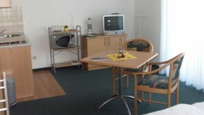 Ein kompaktes Hotelzimmer mit Küchenzeile, einem Einzelbett, einem Esstisch mit Stuhl und einem Fernseher.